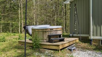 Un bain à remous doté d’une terrasse en bois et d’un petit tabouret, entouré d’une zone boisée.