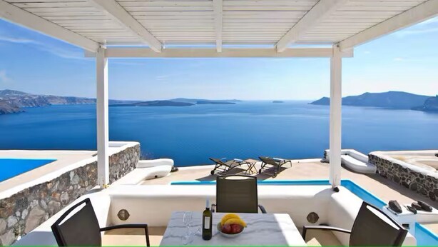 Area salotto esterna coperta con vista sul mare, una piscina e un tavolo con una bottiglia e della frutta.
