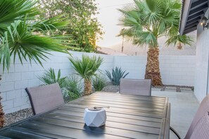 A patio with a table and chairs, surrounded by plants and a white wall. - Cove Pool & Spa · Pets · Single-Level (La Quinta)
