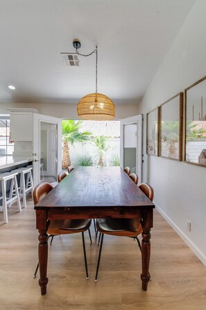 A dining area with a wooden table, chairs, a hanging light fixture, and a view of outdoor plants. - Cove Pool & Spa · Pets · Single-Level (La Quinta)