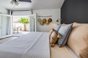 A bedroom with a bed, pillows, a wall-mounted hat rack, and a door leading to an outdoor area with a palm tree. - Cove Pool & Spa · Pets · Single-Level (La Quinta)