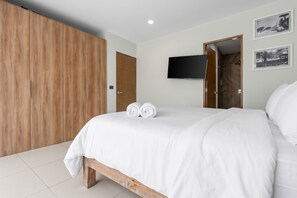 A bedroom with a wooden wardrobe, a bed with white linens, a flat-screen TV mounted on the wall, and two framed pictures. - Stylish Zoho Skies 2-BDR, Rooftop Pool + Parking (Puerto Vallarta)