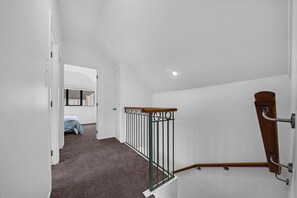 Un couloir doté d’un sol recouvert de moquette, de murs blancs, d’une rambarde en bois et d’une porte donnant sur une pièce où se trouve un lit.