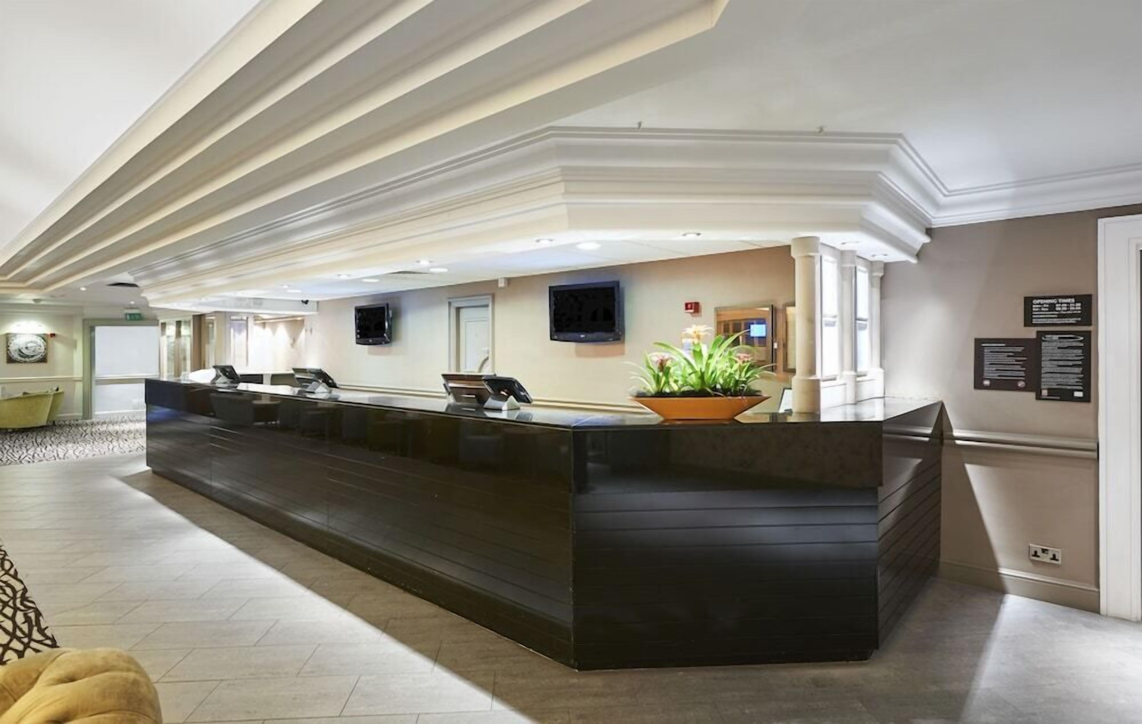 Un área de recepción con un mostrador negro, varias sillas, una planta en maceta, un televisor de pantalla plana y un tablón informativo en la pared.