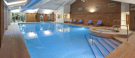 An indoor swimming pool with clear blue water, a sunlit ceiling, and a brick wall.