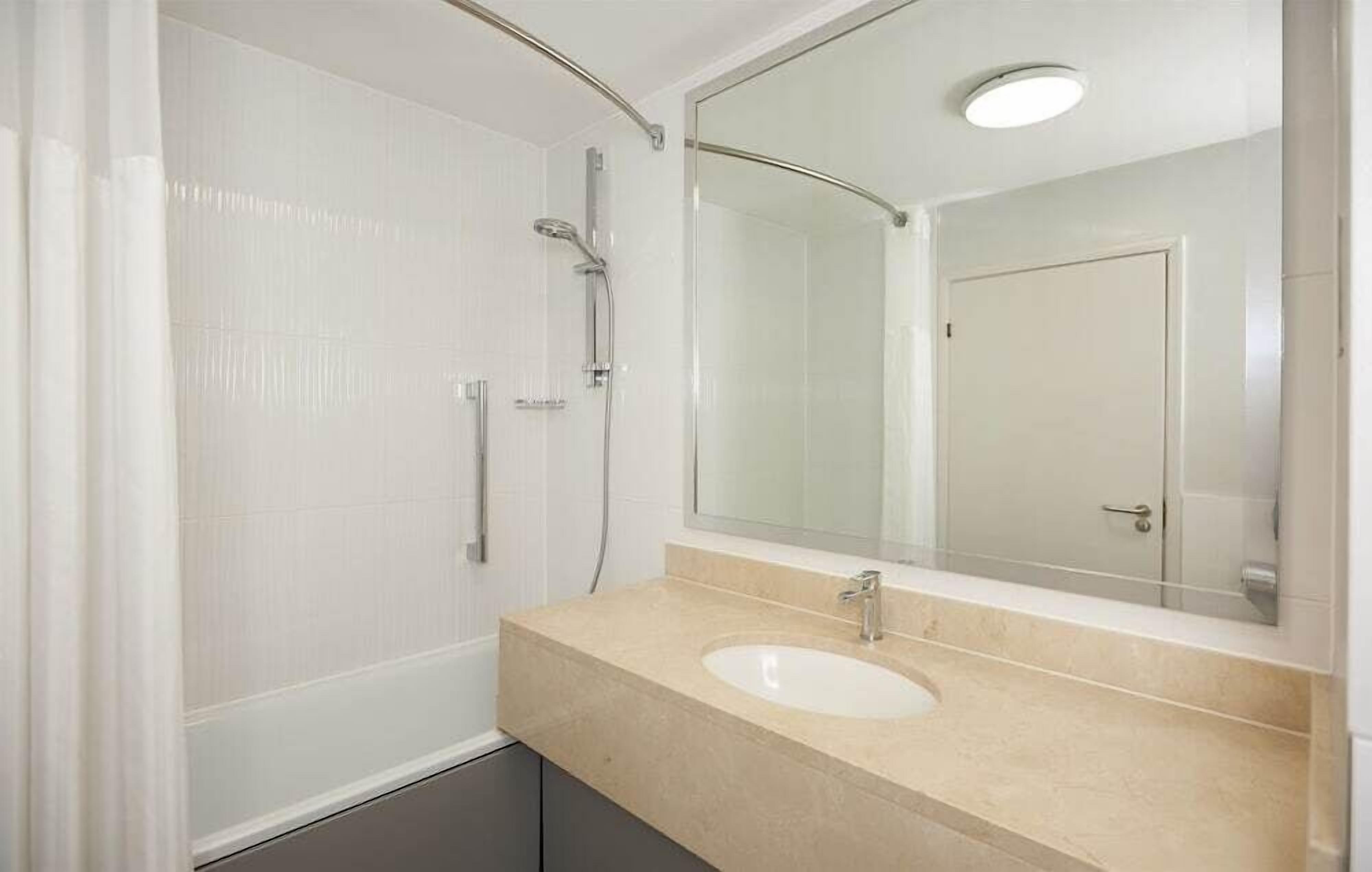 A bathroom with a white tiled wall, a beige countertop, a sink, a mirror, and a shower.