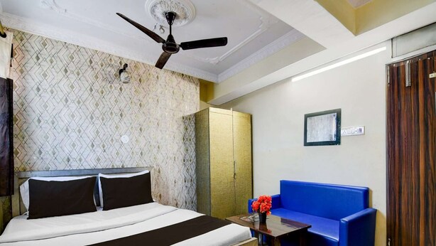 A hotel room with a bed, a blue sofa, a wooden table with a vase of flowers, a ceiling fan, and a wardrobe.