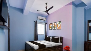 A bedroom with a bed, a ceiling fan, an air conditioning unit, a window with curtains, and a small table with a vase of flowers.