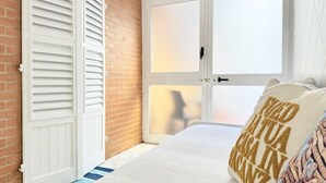 Ein Schlafzimmer mit Backsteinwand, einer weißen Tür mit Fensterläden, einem Bett mit weißer Bettdecke und einem Kissen mit Aufschrift.