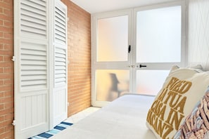 A bedroom with a brick wall, a white door with shutters, a bed with a white comforter, and a pillow with text.