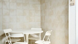 Ein Zimmer mit einem runden Tisch, vier Stühlen, beigefarbenen Fliesenwänden und einer weißen Wand mit strukturierter Oberfläche und einem Holzregal.