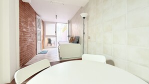 Ein Zimmer mit einem weißen Tisch, zwei weißen Stühlen, einem weißen Sofa, einer Ziegelwand, einem Fliesenboden und einer Tür, die auf einen Balkon führt.