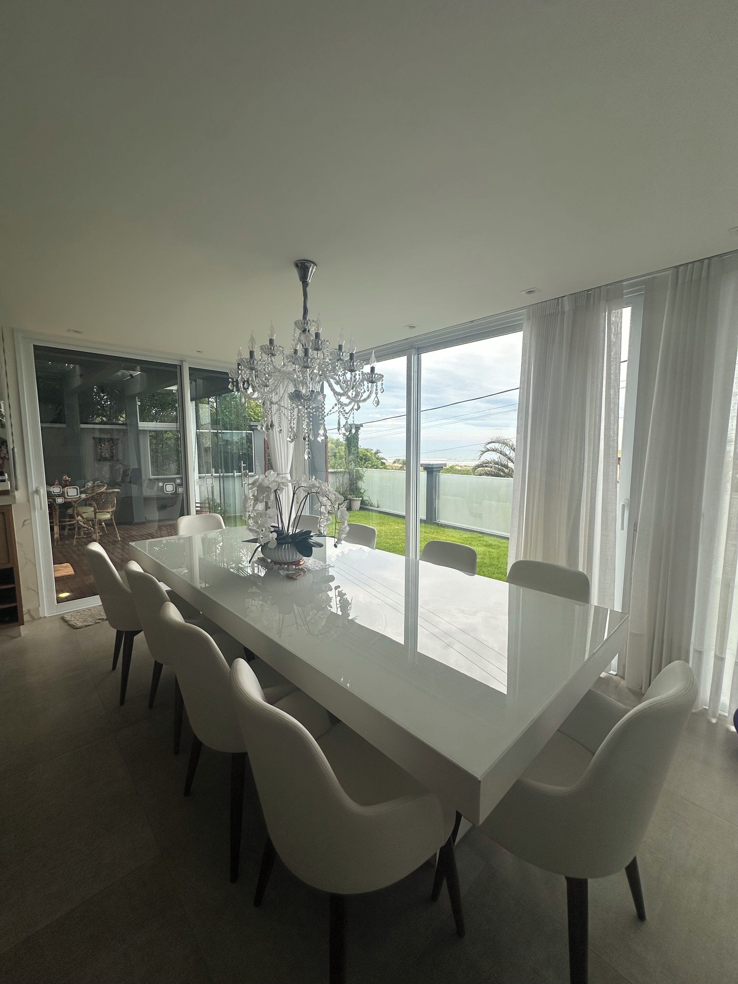 Un comedor con una mesa grande, sillas blancas, una araña y ventanas amplias con cortinas.