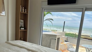 Une chambre avec un grand lit, une étagère en bois, une télévision et une vue sur la plage, accessible par une porte-fenêtre coulissante.
