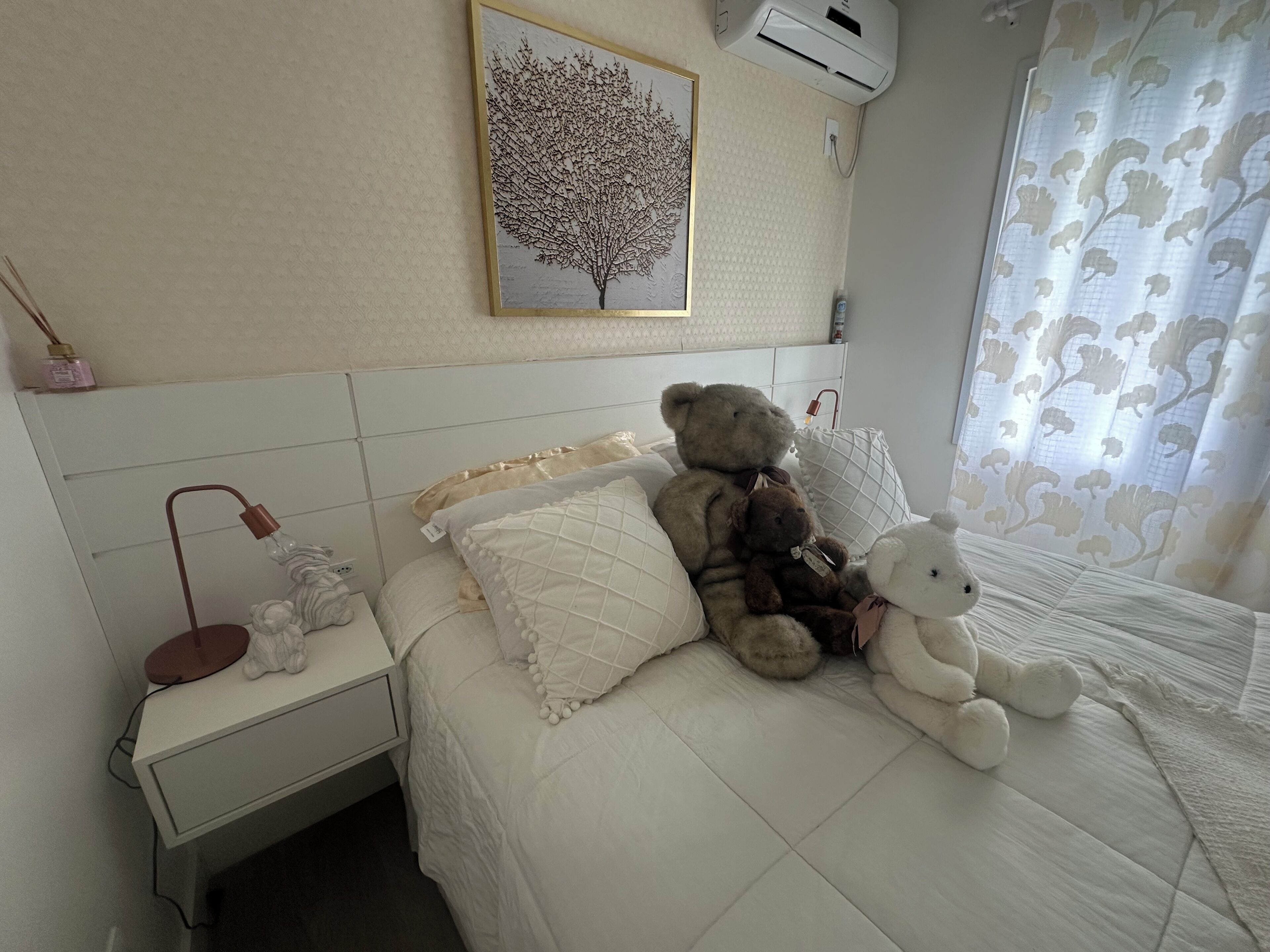 Un dormitorio con cama, almohadas, lámpara de noche, aire acondicionado instalado en la pared, un cuadro enmarco y una ventana con cortinas.