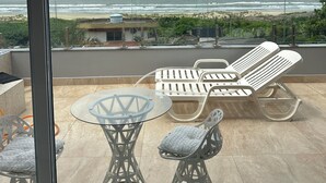 Un balcon avec une table en verre et deux chaises, donnant sur une plage et l’océan.