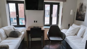 Habitación de hotel con sofá blanco, una silla negra, un escritorio, un televisor de pantalla plana y ventanas amplias con vistas a edificios.