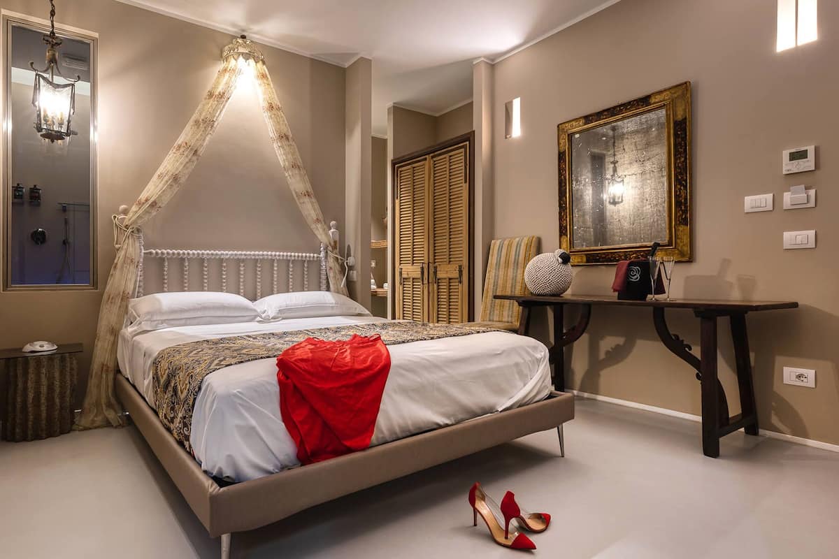 A bedroom with a canopy bed, a bedside table, a mirror, and a wall-mounted thermostat.