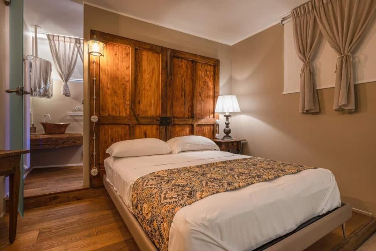 A bedroom with a wooden headboard, a bedside table with a lamp, a mirror, and a window with curtains.