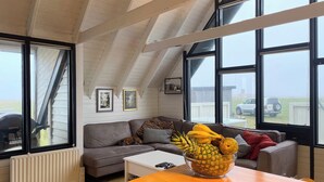 Ein gemütliches Wohnzimmer mit Holzbalkendecke, einem grauen Sofa, einem weißen Couchtisch und einer Obstschale auf dem Tisch.