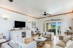 A spacious living area with a TV, white cabinetry, and a view through an arched doorway to an outdoor patio. - Sugar Hill Birds Eye Villa - by Zenbreak (Holetown)