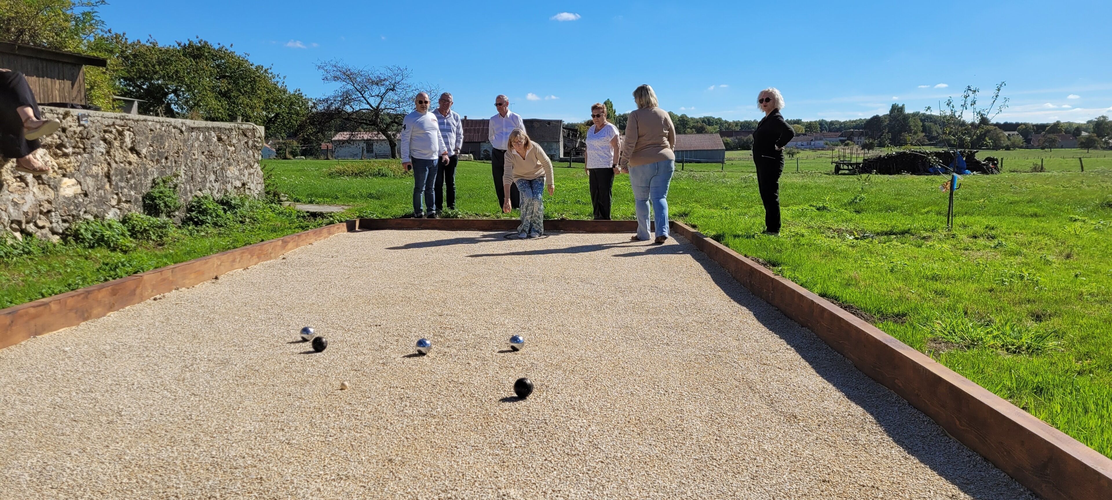A group of people standing around a croquet field with a stone wall and a wooden fence in the background.