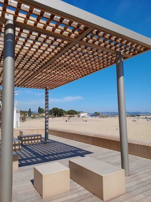 Ein überdachter Außenbereich mit Holzlattendecke, Betonbänken und Blick auf einen Strand.