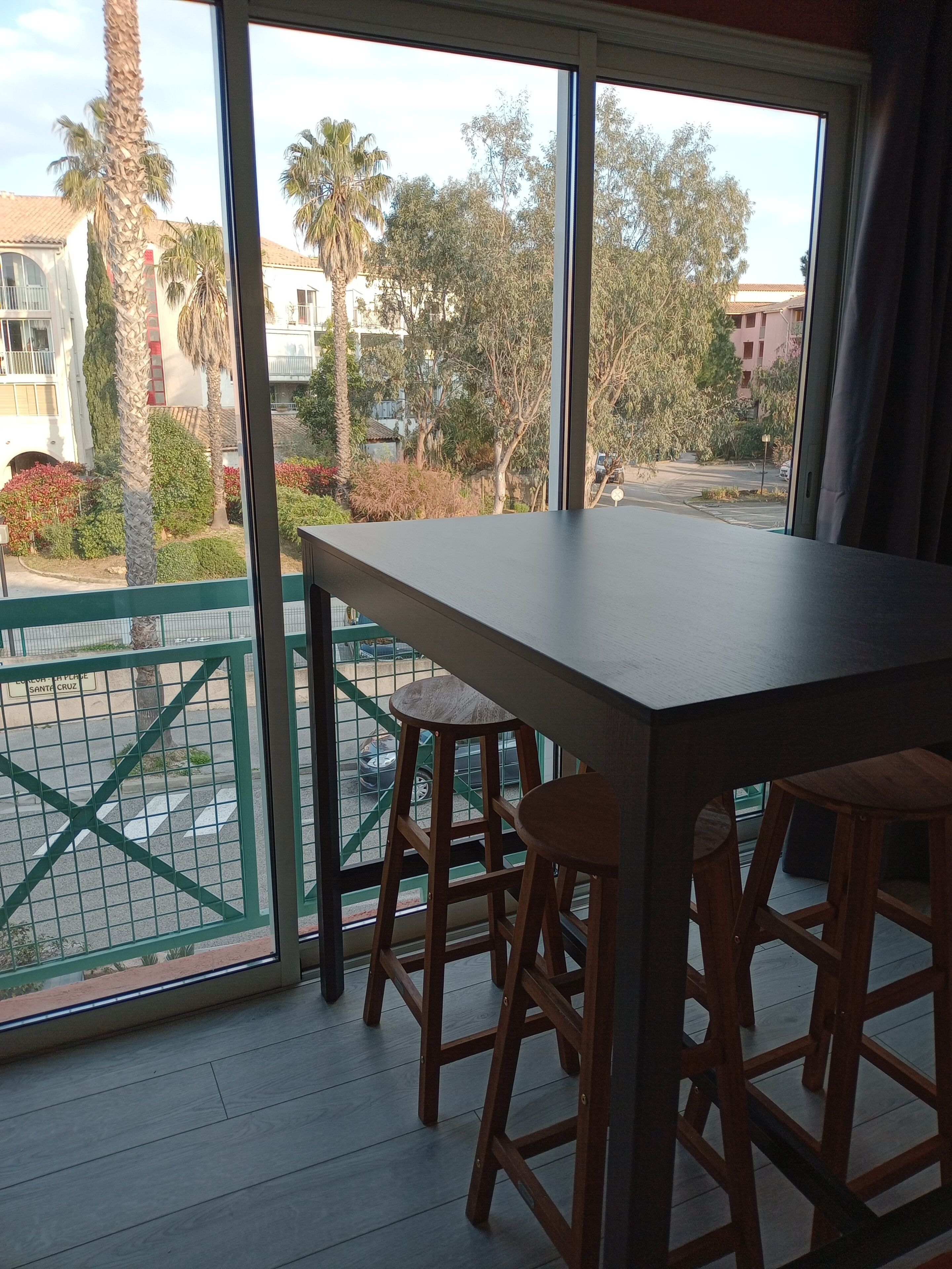 A balcony with a table and stools, a view of a street with palm trees and buildings, and a green metal railing.
