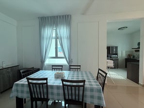 Un coin repas avec une nappe à carreaux, des chaises en bois, une fenêtre avec des rideaux et, au fond, une cuisine visible.