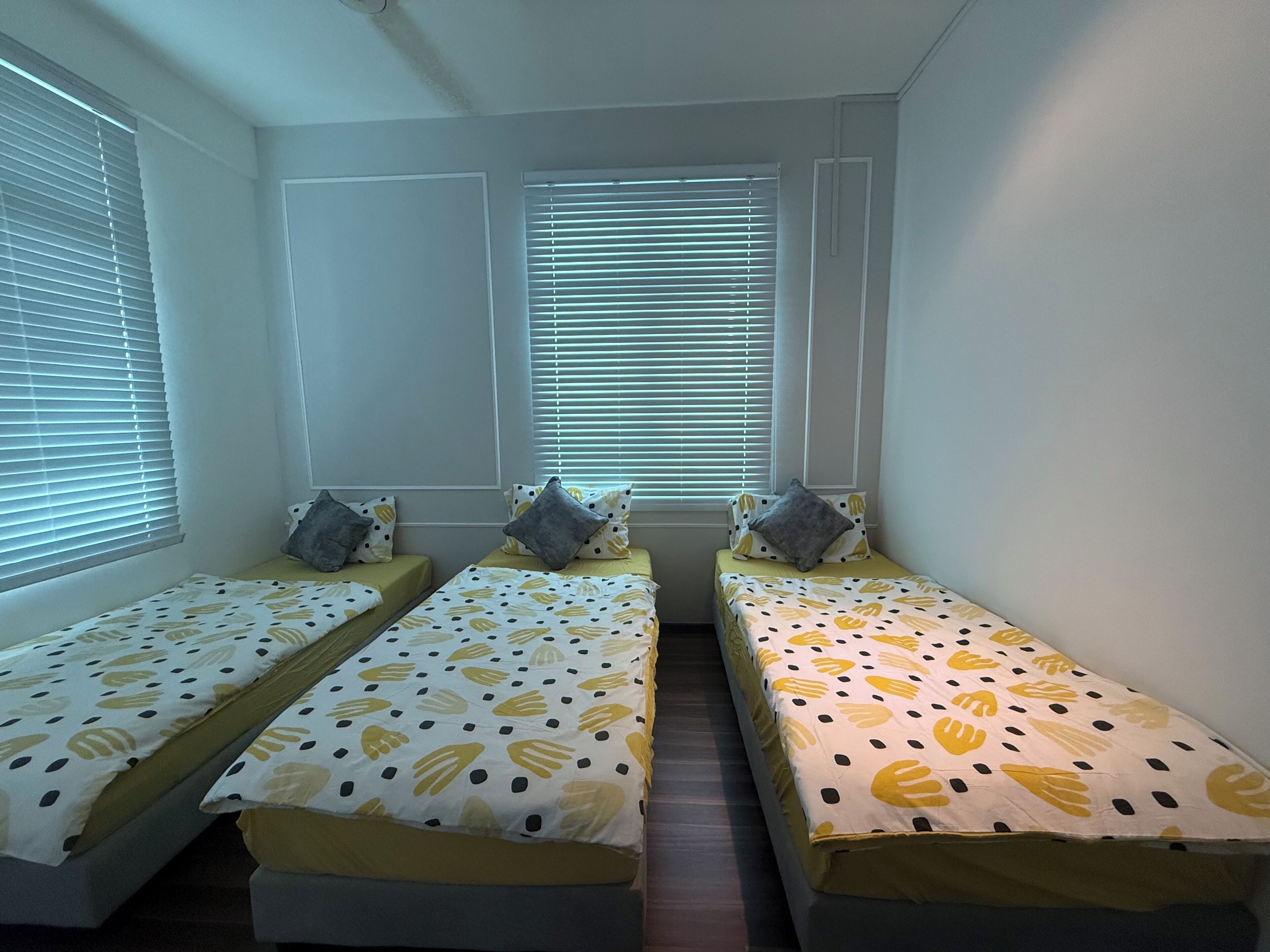 Quarto com três camas individuais, cada uma com colcha e almofadas estampadas, e uma janela com persianas.