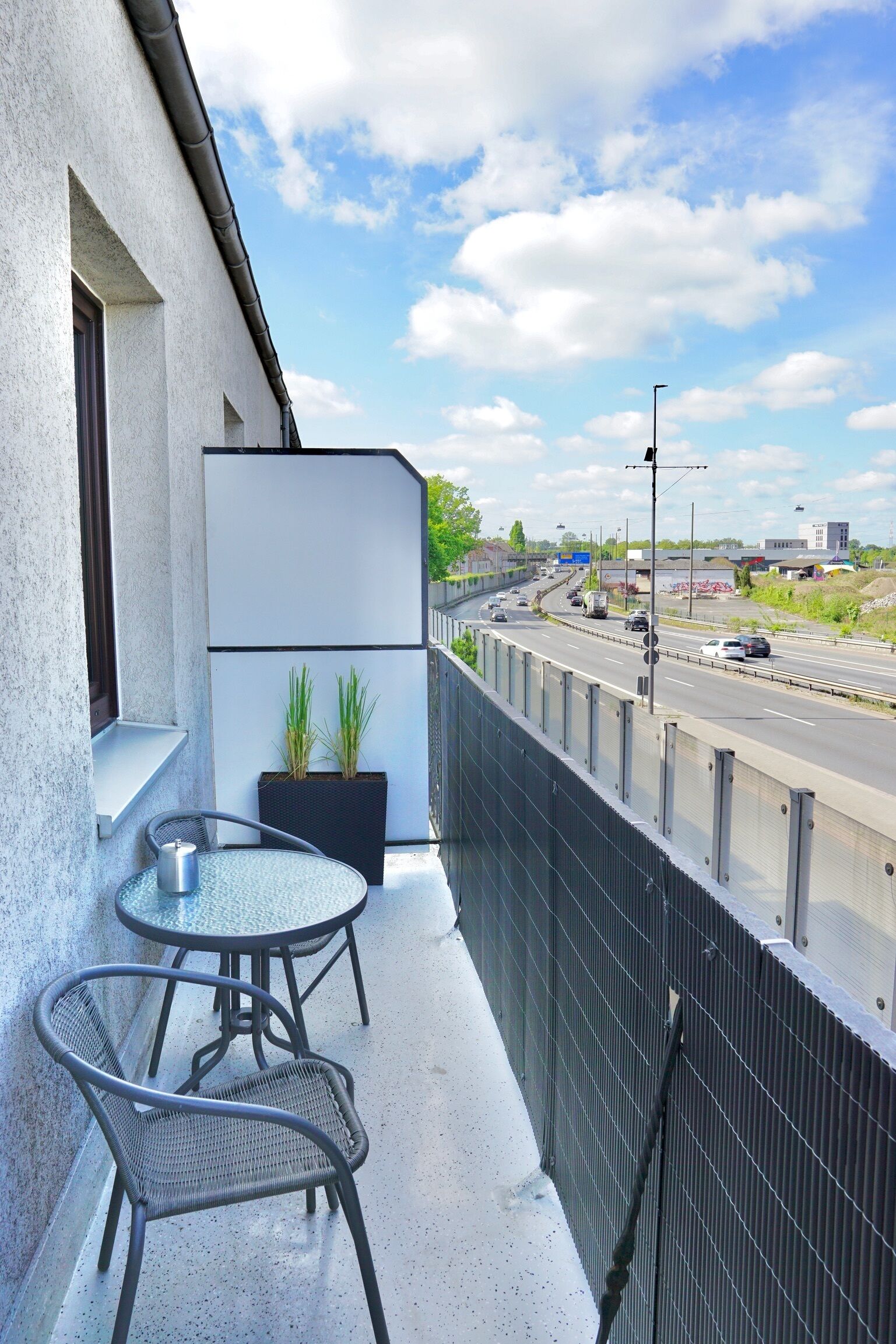 Ein Balkon mit Glastisch und Stühlen, einem Blumentopf und Blick auf eine Autobahn.