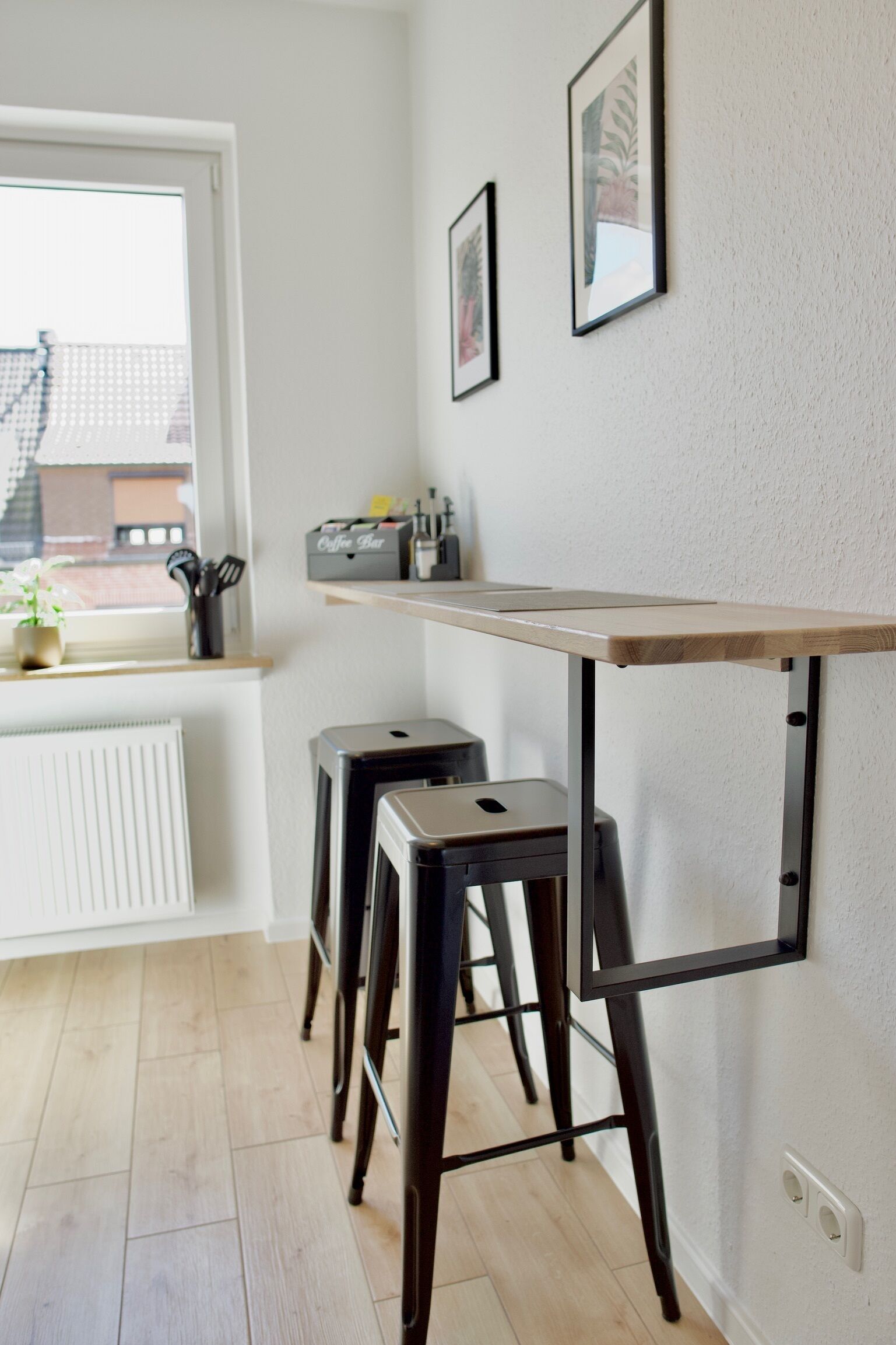Eine Küche mit einem Holztisch, zwei schwarzen Hocker, einem Heizkörper und einem Fenster mit Blick auf Gebäude.