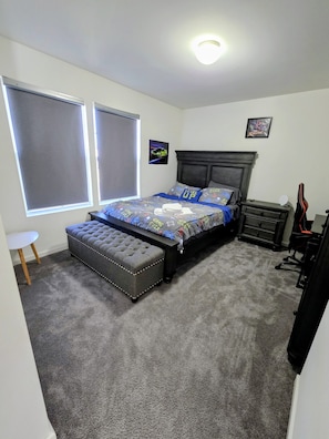 Une chambre à coucher comprenant un lit, un bureau, une chaise, une fenêtre avec des stores, un tableau accroché au mur et un luminaire au plafond.