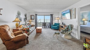 A living room with a sofa, armchair, and ottoman, a blue armchair, a small table with a lamp, and a bed with a view. - 🌊 🏖️ Oasis 7: Walk to Beach, Private Patio + Perfect Daytona Base! (Daytona Beach Shores)