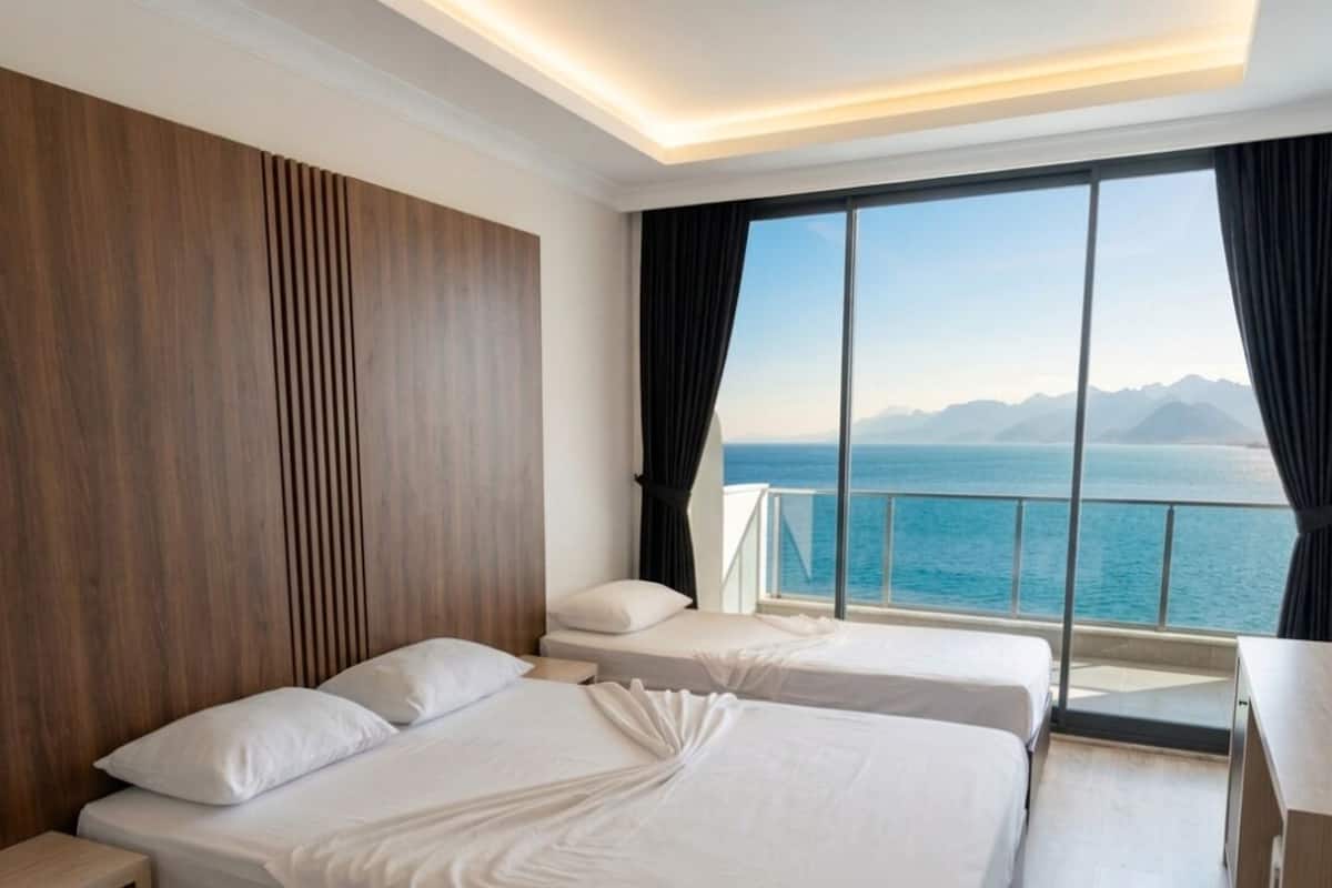 Habitación de hotel con dos camas, un ventanal con vistas al mar y a las montañas, y cabecero de madera.