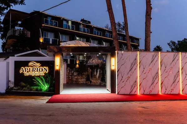 A hotel entrance with a sign reading 'APERION BEACH HOTEL,' a red carpet, and a stone wall with a statue. - Club Aperion Beach Hotel (Manavgat)