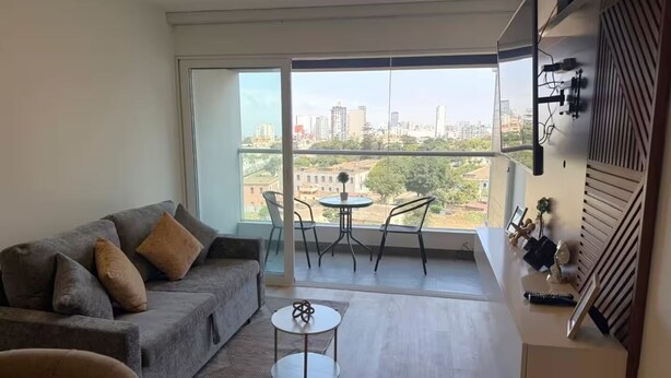 En moderne lejlighed med en grå sofa, et lille rundt bord og en balkon med udsigt over byen.