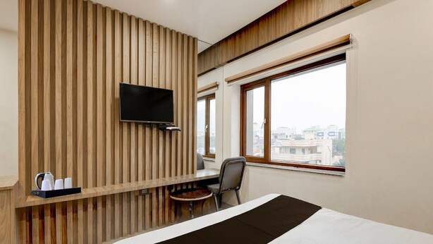 Une chambre d’hôtel comprenant un lit, un téléviseur fixé sur un mur en panneaux de bois, une petite table avec une bouilloire et des tasses, une chaise et une fenêtre donnant sur la ville.