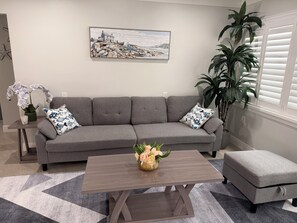 Ein Wohnzimmer mit grauem Sofa, einem Couchtisch mit Blumen, einem Bild an der Wand und einer Topfpflanze.