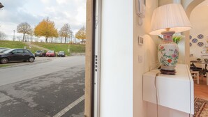 Huoneessa on valkoinen konsolipöytä, koristeellinen lamppu ja ikkunasta avautuu näkymä parkeeraatuihin autoihin ja puihin.