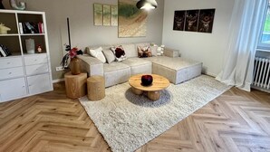 Una sala de estar moderna con un sofá modular, una mesa de centro redonda de madera, una estantería blanca y un amplio ventanal con cortinas.