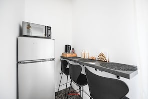 Uma cozinha compacta equipada com frigorífico, micro-ondas, máquina de café e uma pequena área de refeições com cadeiras e mesa.