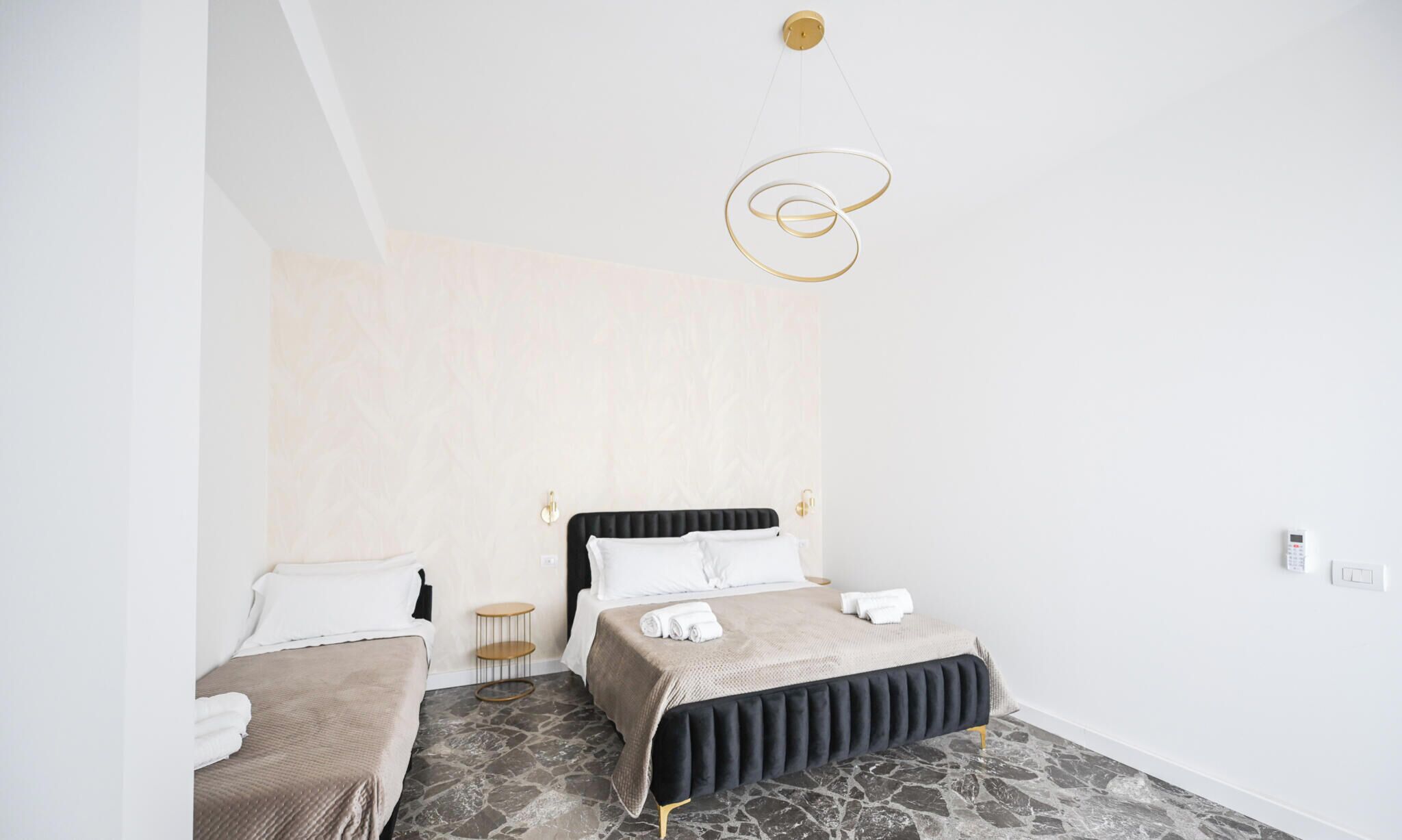 A bedroom with two beds, a stone-patterned floor, a hanging light fixture, and a plain wall.