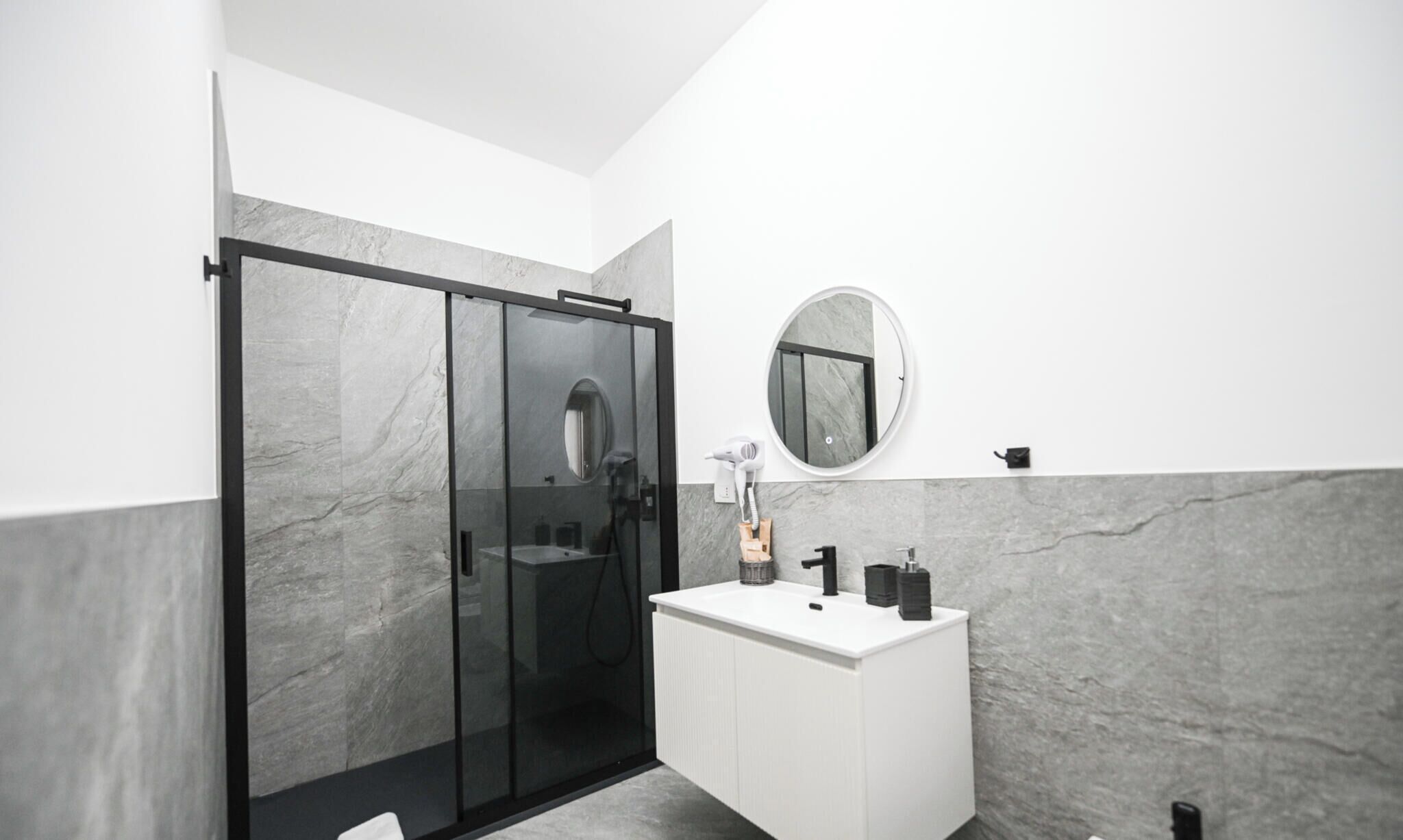 A modern bathroom with a glass-enclosed shower, a white vanity with a round mirror, and marble walls.