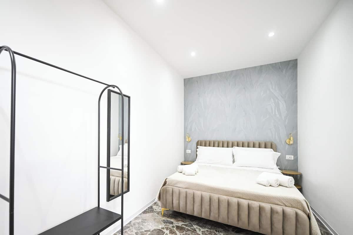 A modern bedroom with a bed, a bedside table, a wall-mounted mirror, and a control panel.