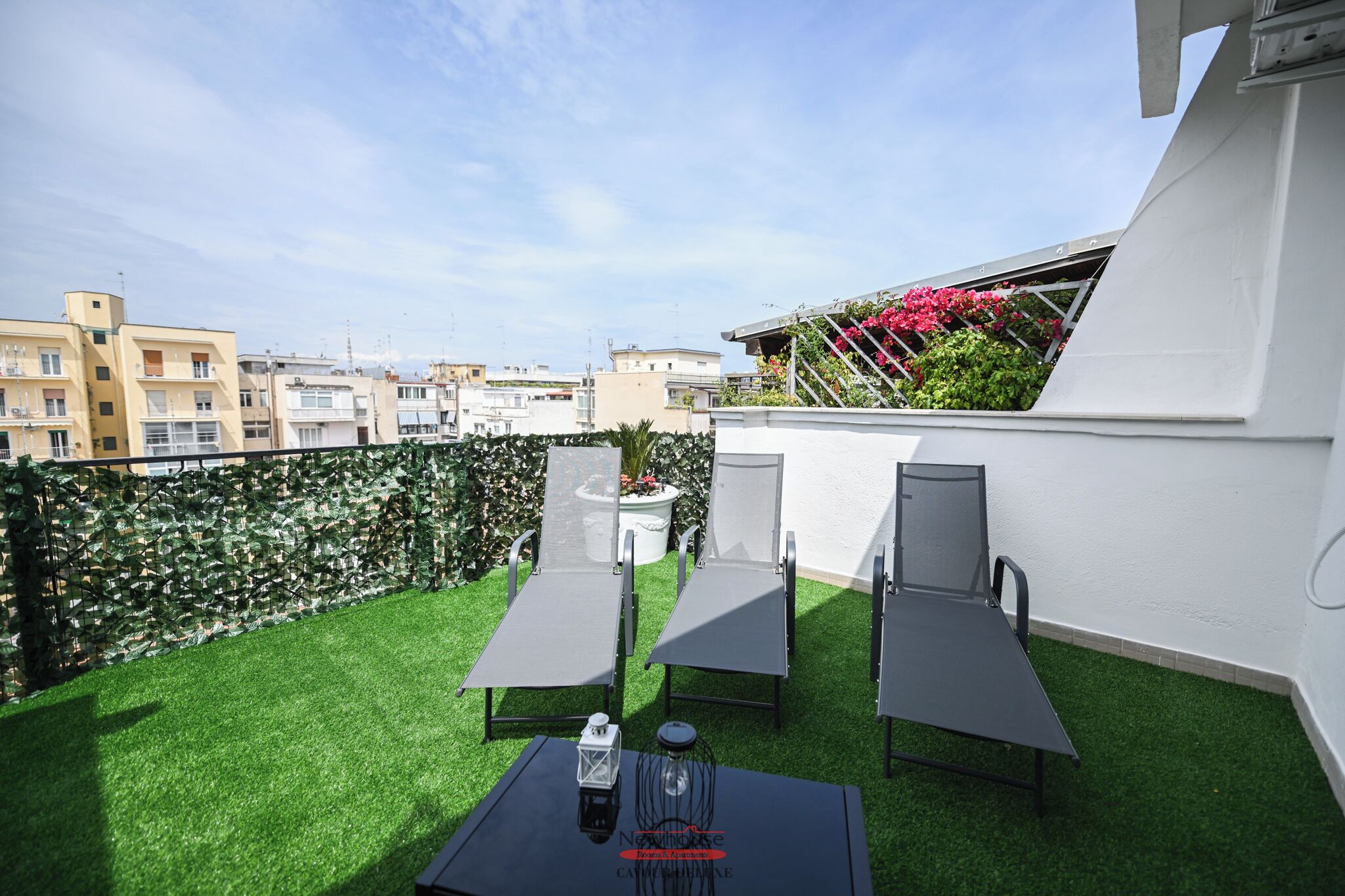 Een dakterras met kunstgras, twee loungesteilen, een tafel met een fles en lantaarn, en een witte muur met een balkon.