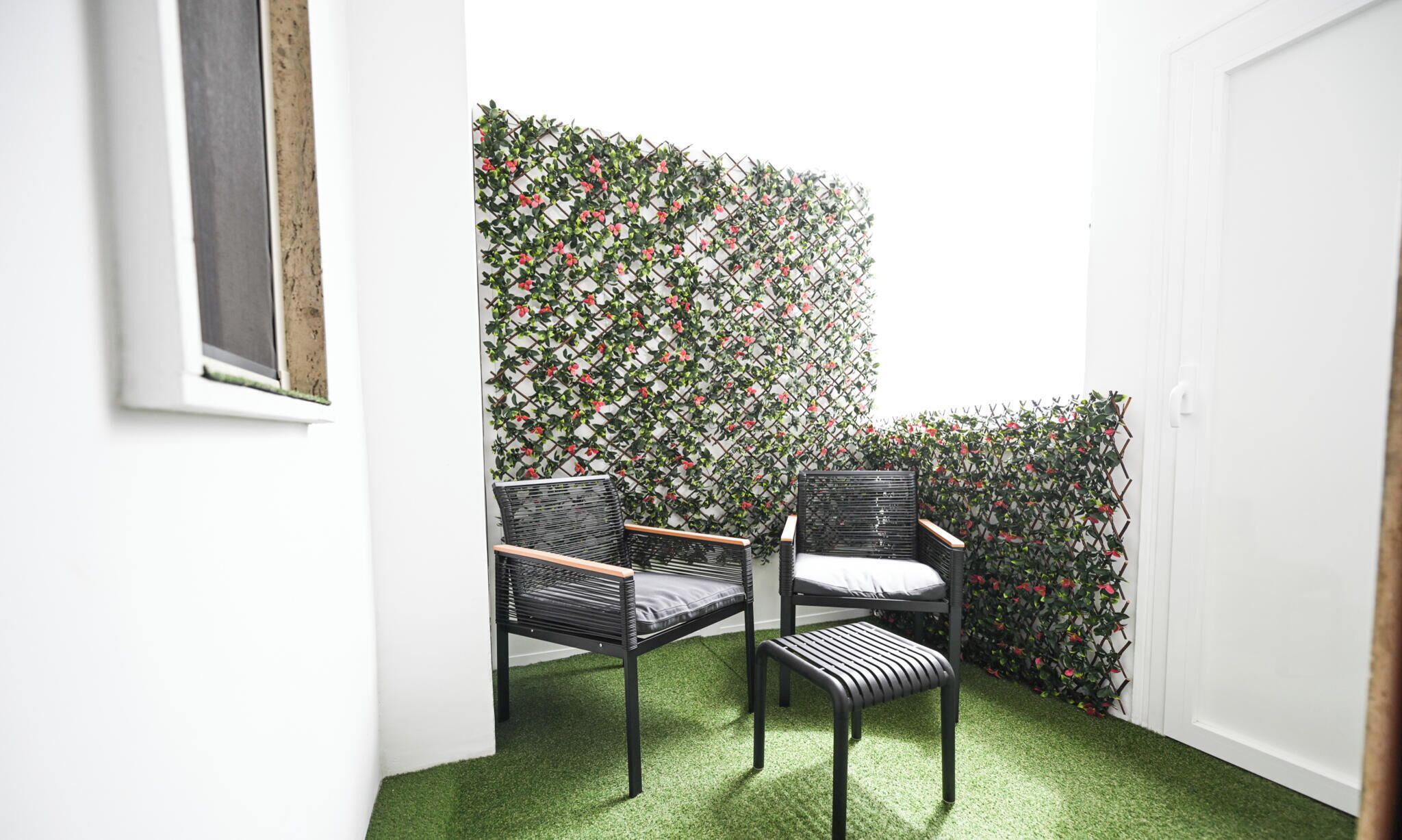 Habitación de esquina con piso de césped artificial verde, dos sillas de mimbre negro y una pared decorativa con un patrón de celosía y flores rojas.