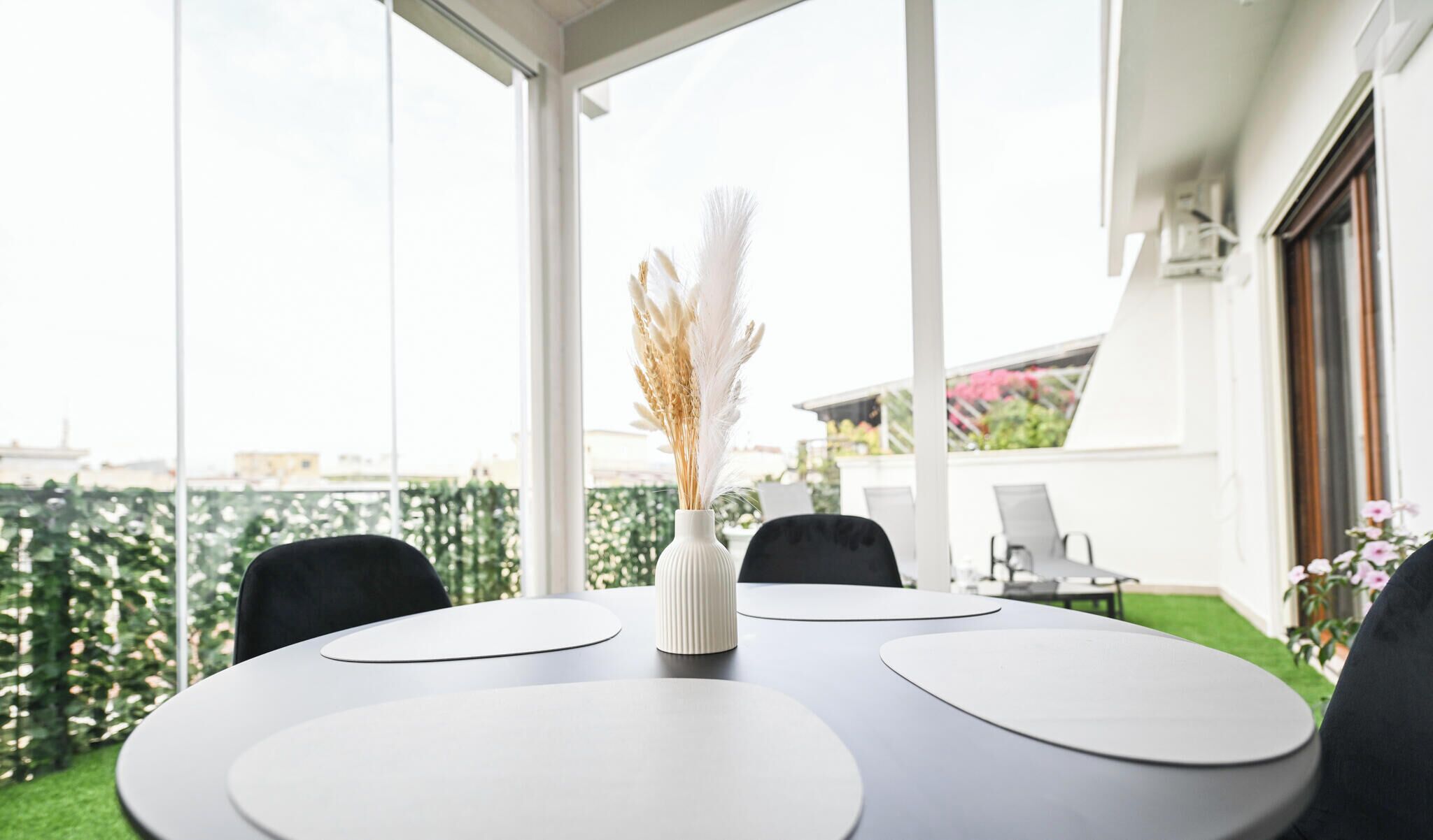 Un área de comedor al aire libre moderna, con una mesa y sillas blancas, un florero con plumas blancas y vistas a un paisaje urbano a través de amplias ventanas.