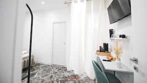 Habitación de hotel moderna con suelo de diseño de piedra, cama con cabecera blanca, escritorio con silla, televisor montado y zona de ducha con cortinas blancas.