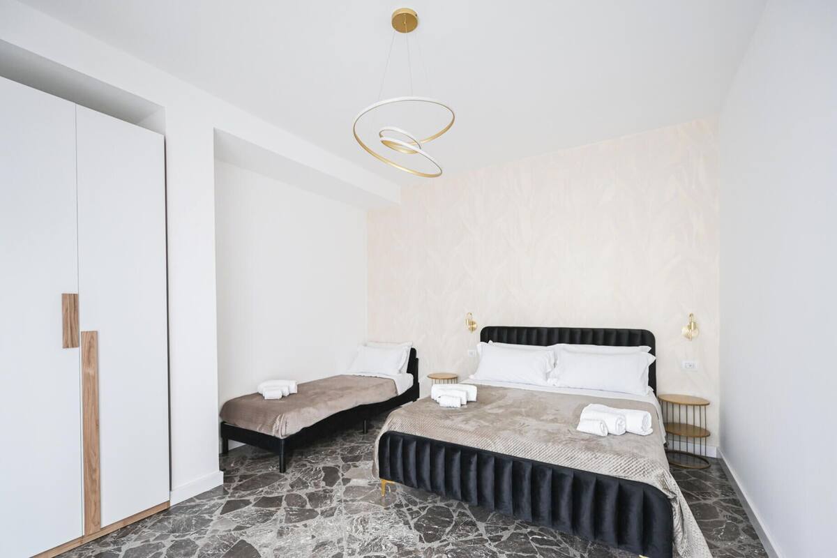A bedroom with a large bed, a smaller bed, a wardrobe, a nightstand, and a modern light fixture.